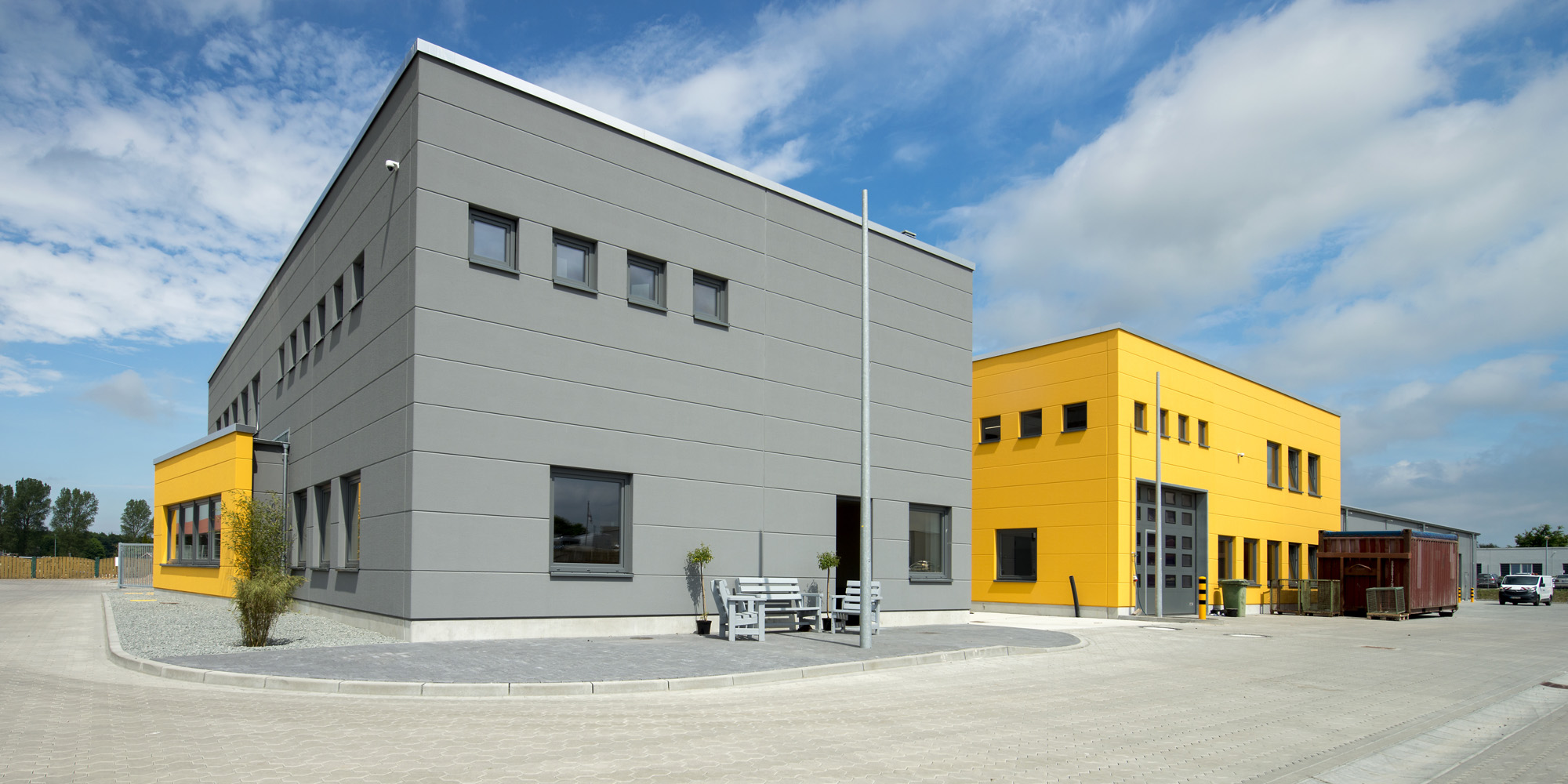 Ein mehrstöckiger Betriebshof mit einladendem Vorplatz, die graue Fassade aus Porenbeton wird von leuchtend gelben Elementen durchbrochen